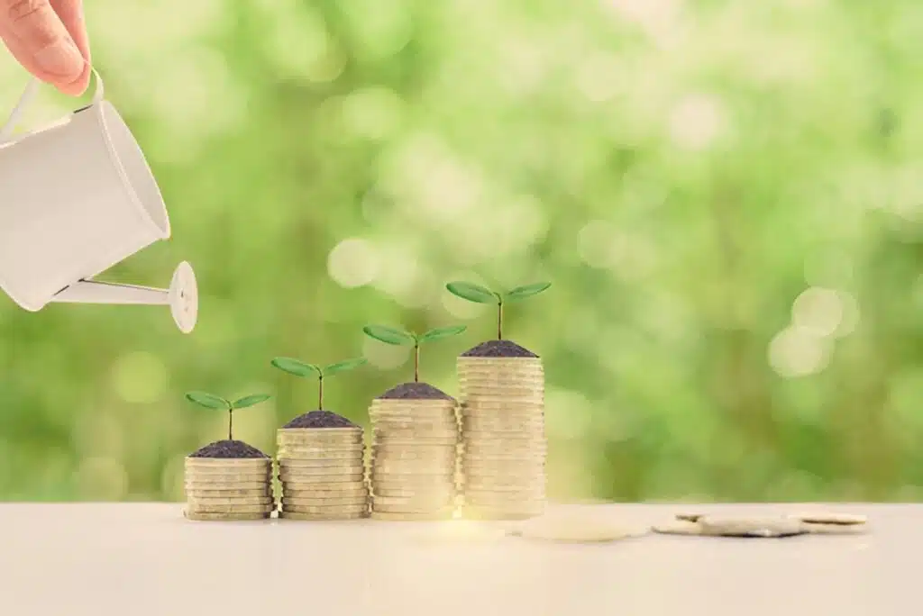 A white watering can pouring water over four ascending stacks of coins with small green seedlings growing from soil on top of each stack, against a blurred green background